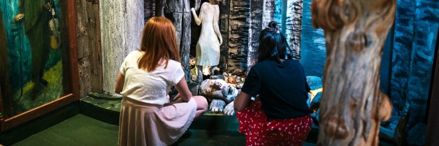 Two girls are visiting the museum about the storytelling network in Kronoberg and are in a room that is set up as a forest.